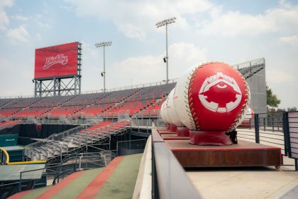 Estádio de beisebol vazio, com grandes elementos decorativos alusivos ao esporte, sob céu claro.
